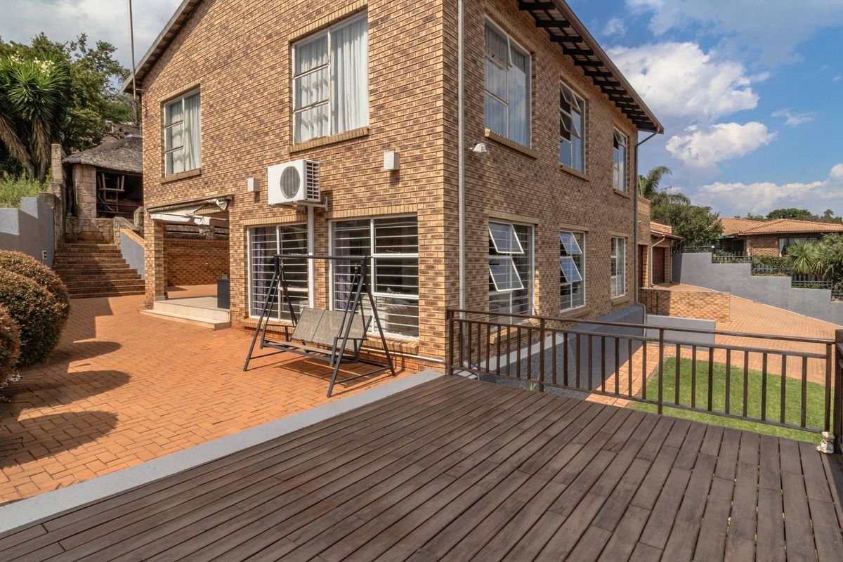 A view of the rear exterior of a double-story brick house featuring a wooden deck, paved patio area, and landscaped garden surroundings.
