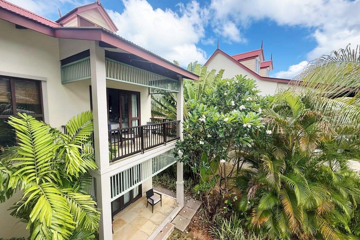 A two-story maison de ville featuring a covered balcony and ground-level patio, surrounded by lush tropical landscaping and mature trees.