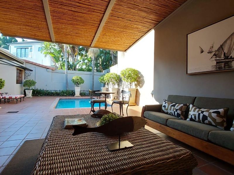 A spacious covered patio featuring bamboo ceiling detail and tiled flooring, overlooking a private swimming pool and landscaped garden area.
