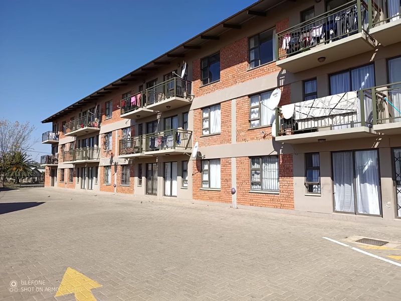 Exterior view of a multi-story apartment complex featuring brick facade, private balconies with metal railings, and a paved parking area.