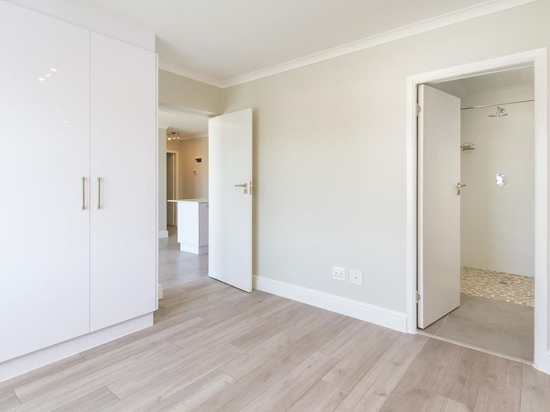 A bright, modern bedroom featuring light laminate flooring, built-in white cupboards, and direct access to an en-suite bathroom.