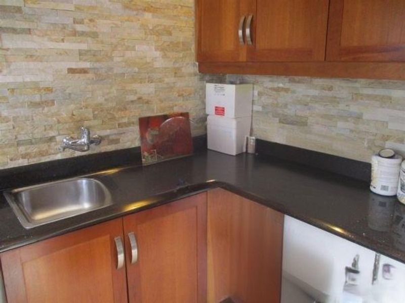 A kitchen corner featuring wooden cabinetry, dark granite countertops, a stainless steel sink, and a textured stone veneer backsplash.