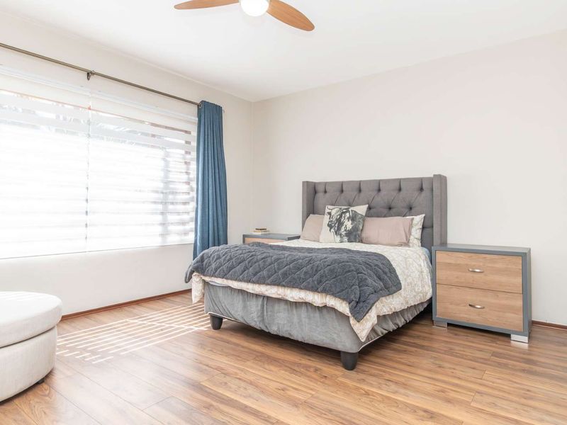 A well-lit bedroom featuring durable laminate flooring, a ceiling fan, and large windows with venetian blinds for natural light control.