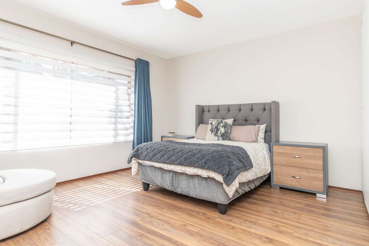 A well-lit bedroom featuring durable laminate flooring, a ceiling fan, and large windows with venetian blinds for natural light control.