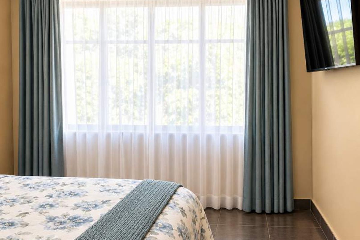 A well-maintained bedroom featuring exposed timber ceiling beams, tiled flooring, and a large window with floor-to-ceiling curtains.