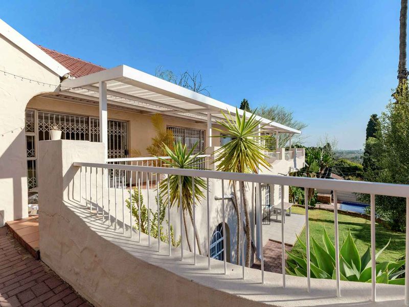 A spacious covered patio featuring a metal balustrade, tiled flooring, and security gates, overlooking the garden and pool area.