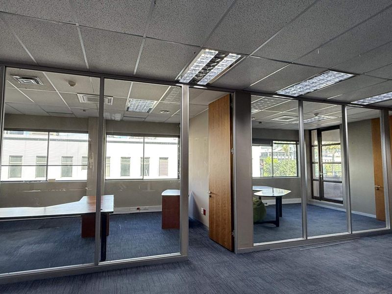 Open-plan office space featuring glass partitions, suspended ceilings with recessed lighting, and large windows providing natural light.