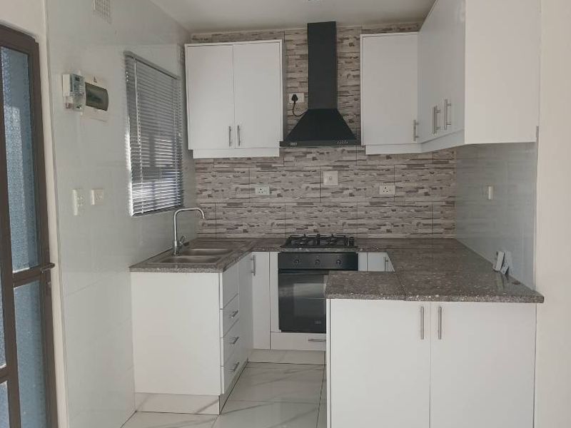 A clean, modern kitchen featuring white cabinetry, stone countertops, a gas hob with extractor fan, and light-colored tiled flooring throughout.