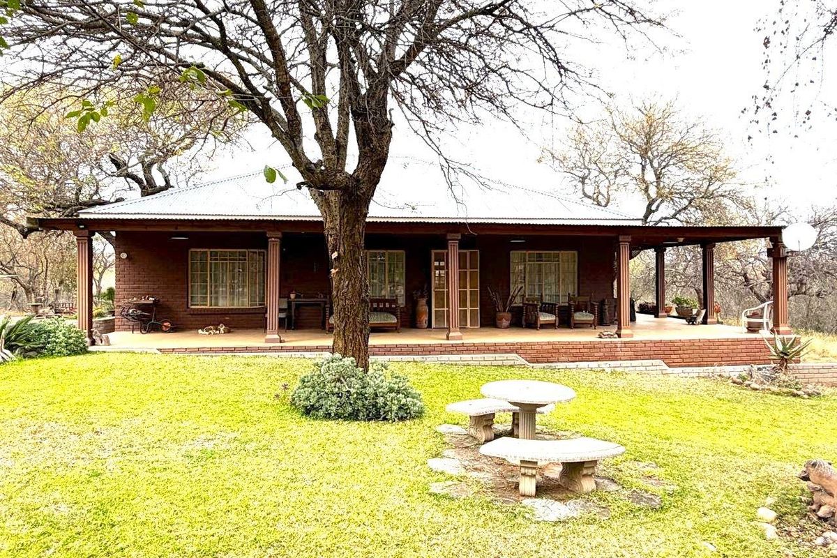 A charming single-story home featuring a spacious covered patio, brick exterior, and a metal roof, set within a mature, tree-lined garden.