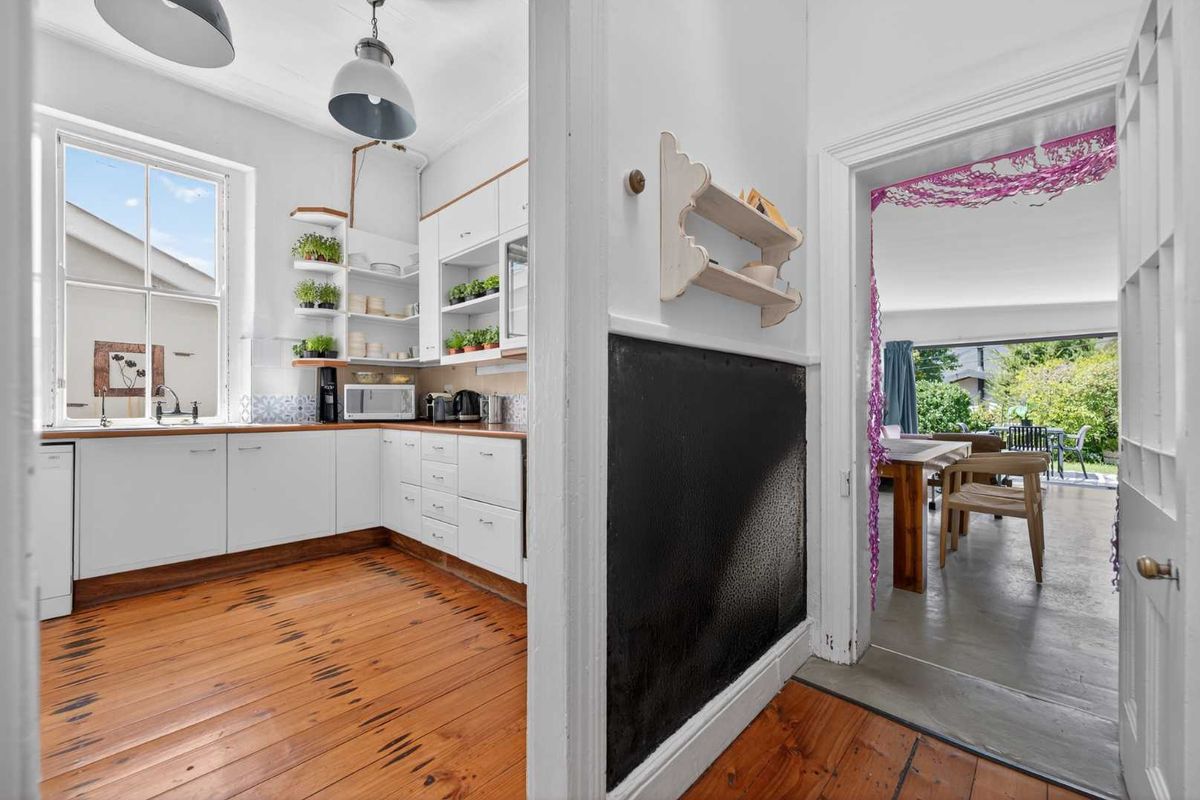 A bright kitchen featuring timber flooring, white cabinetry, and a sash window, with an adjacent view through to the dining area and garden.