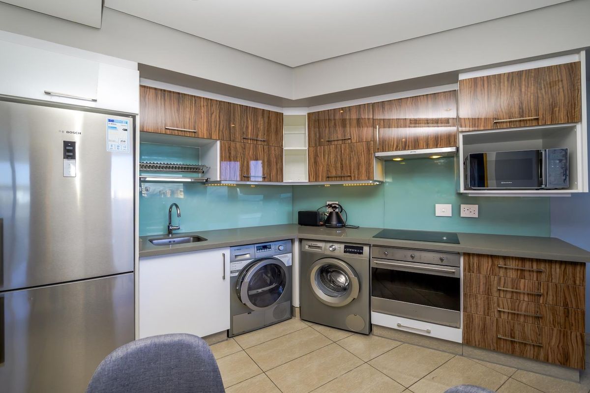Modern kitchen featuring wood-grain cabinetry, stone countertops, glass splashback, and integrated stainless steel appliances including an oven, cooktop, and microwave.