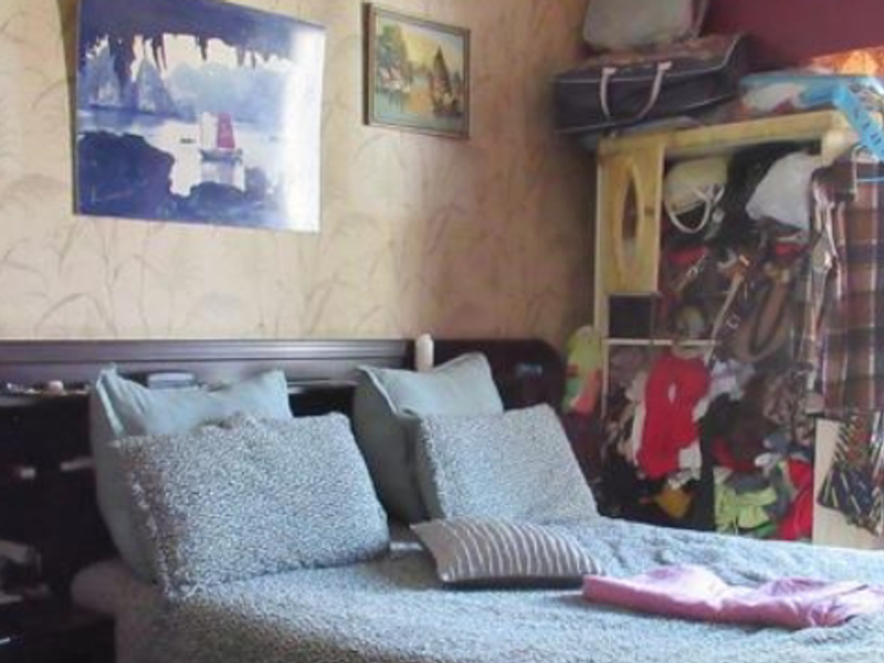 A bedroom featuring a built-in headboard and patterned wallpaper, currently cluttered with personal belongings.