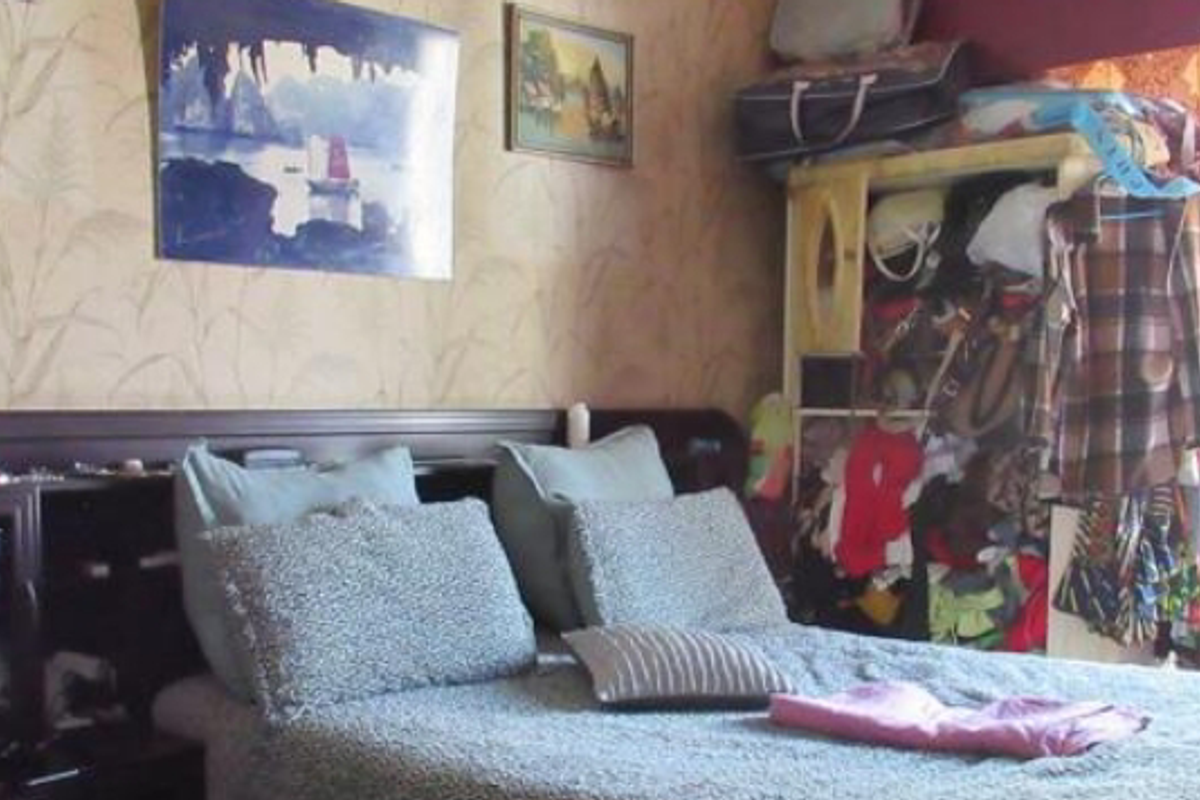 A bedroom featuring a built-in headboard and patterned wallpaper, currently cluttered with personal belongings.