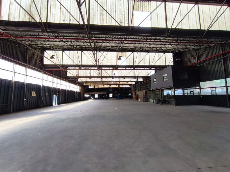 Spacious industrial warehouse featuring high ceilings with exposed steel trusses, translucent roof panels for natural light, concrete flooring, and an integrated mezzanine office structure.