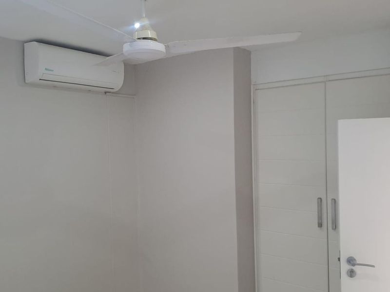 A clean, neutral-toned bedroom featuring a built-in wardrobe, ceiling fan, and wall-mounted air conditioning unit.