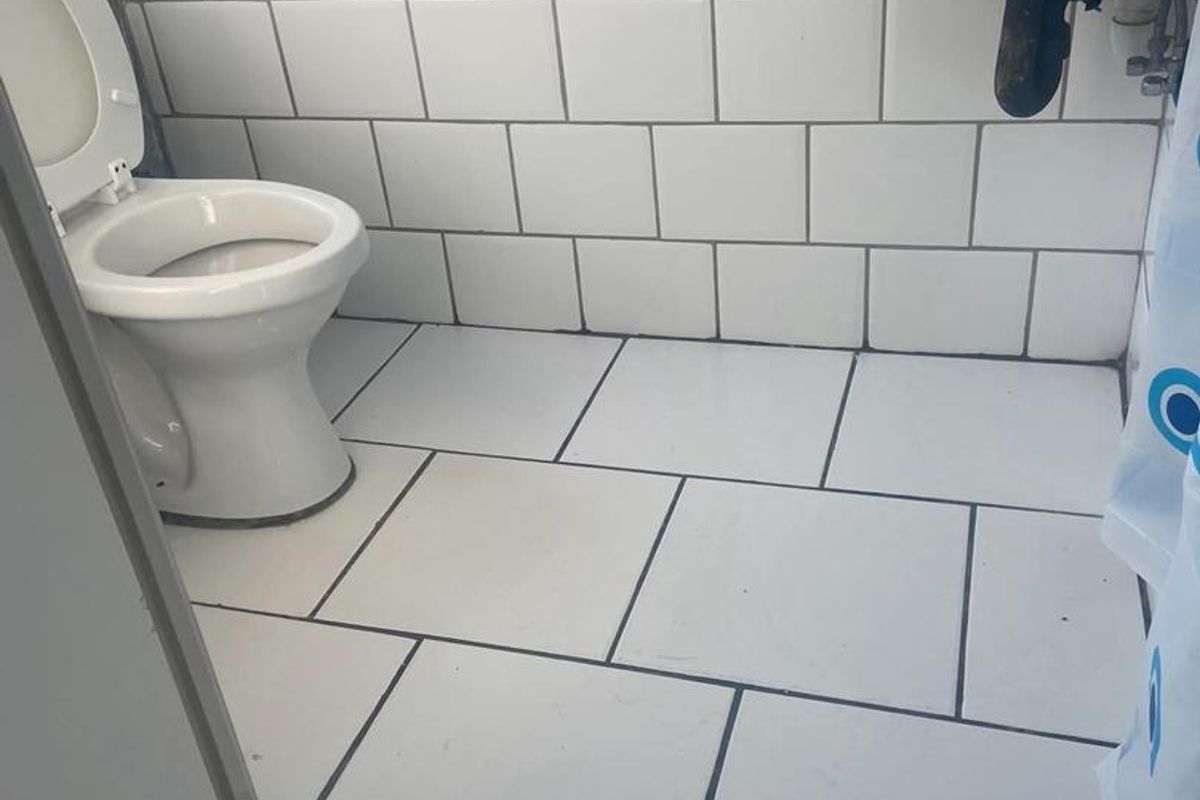A compact bathroom featuring white wall and floor tiles, a standard toilet, and a wall-mounted basin with exposed plumbing.
