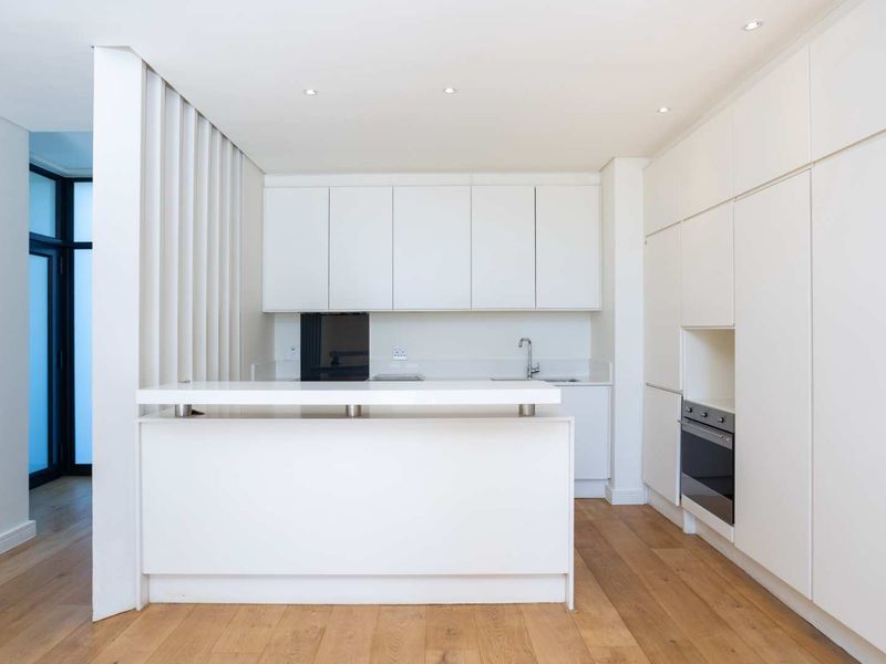 Modern minimalist kitchen featuring sleek white cabinetry, integrated oven, stone countertops, and warm timber flooring in an open-plan layout.