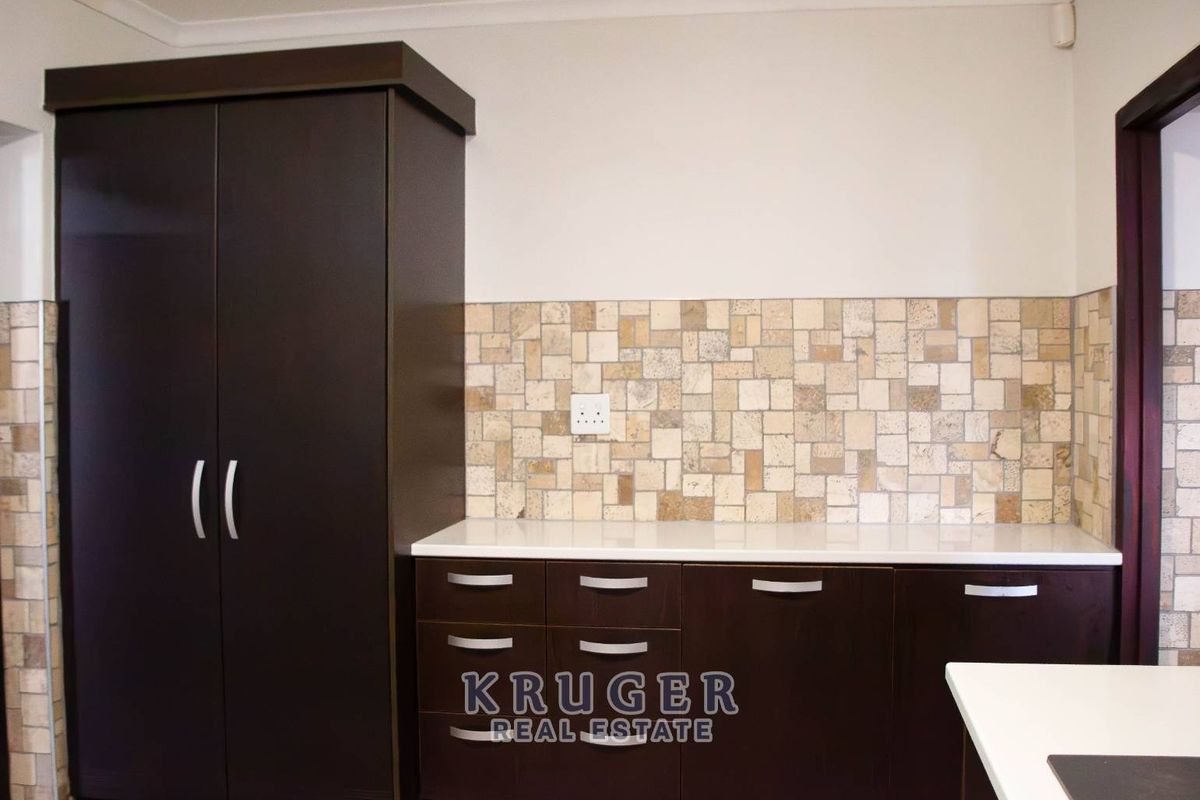 A clean kitchen area featuring dark wood-finish cabinetry, a light stone mosaic backsplash, and white countertops, providing a functional and neutral prep space.