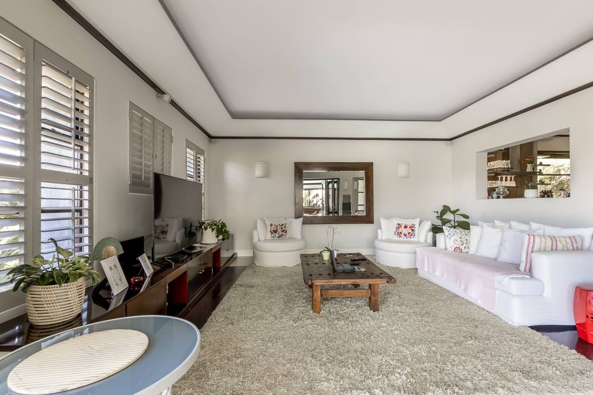 A bright and spacious living room featuring recessed tray ceilings, plantation shutters, and a convenient pass-through window to the adjacent kitchen area.