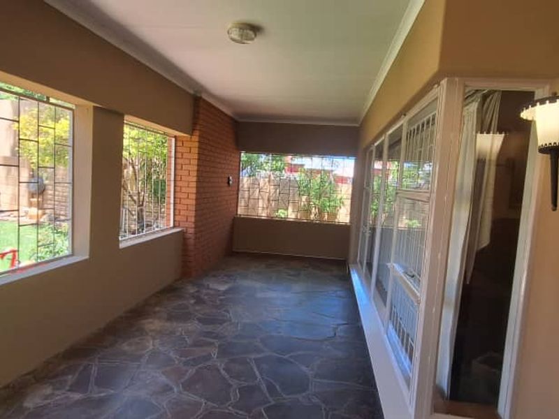 A covered patio area featuring stone flooring, exposed brick accents, and large windows with security bars, offering a view of the garden.
