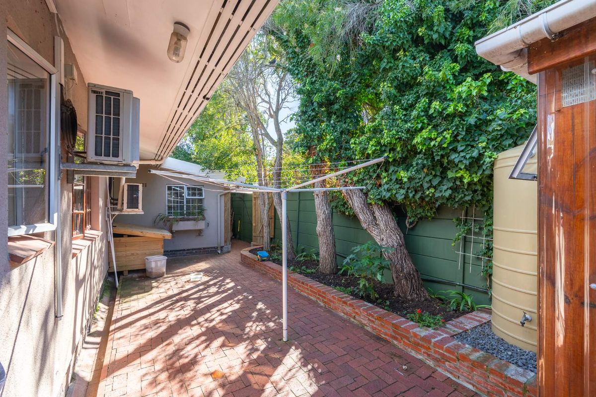 A paved side courtyard featuring a water storage tank, wall-mounted air conditioning unit, and established garden borders with mature trees.