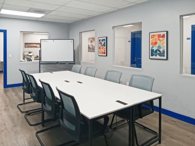 A modern, well-lit office meeting room featuring laminate flooring, internal glass partitions, and a professional conference setup with integrated power outlets.