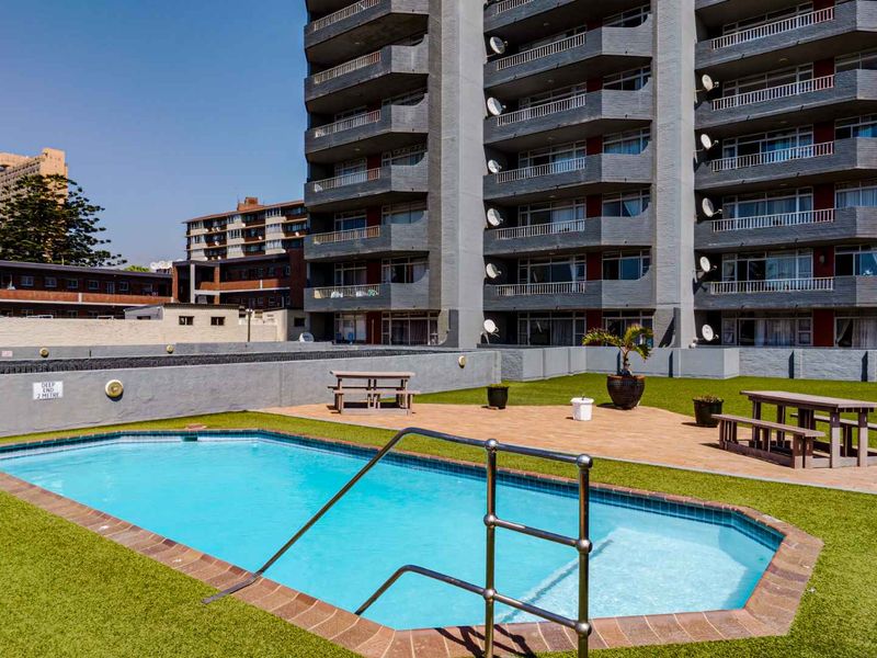 Communal outdoor pool area featuring a swimming pool, paved patio space, and artificial lawn set against the apartment building facade.