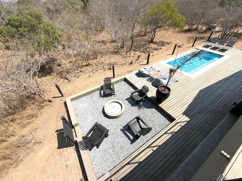 An elevated view of an outdoor entertainment area featuring a swimming pool, timber decking, and a sunken fire pit area set in a natural landscape.