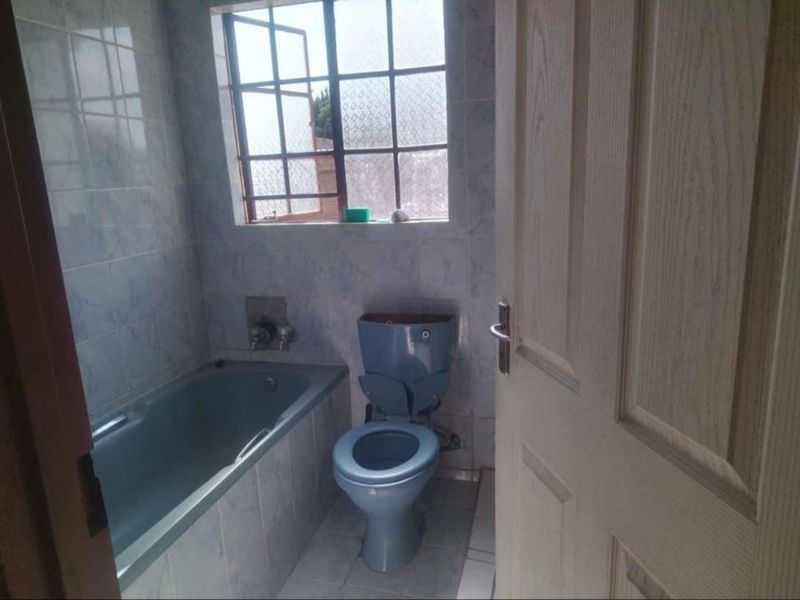 A dated bathroom featuring a blue bathtub and toilet, tiled surfaces, and a window with security bars. Requires modernization and repair of fixtures.