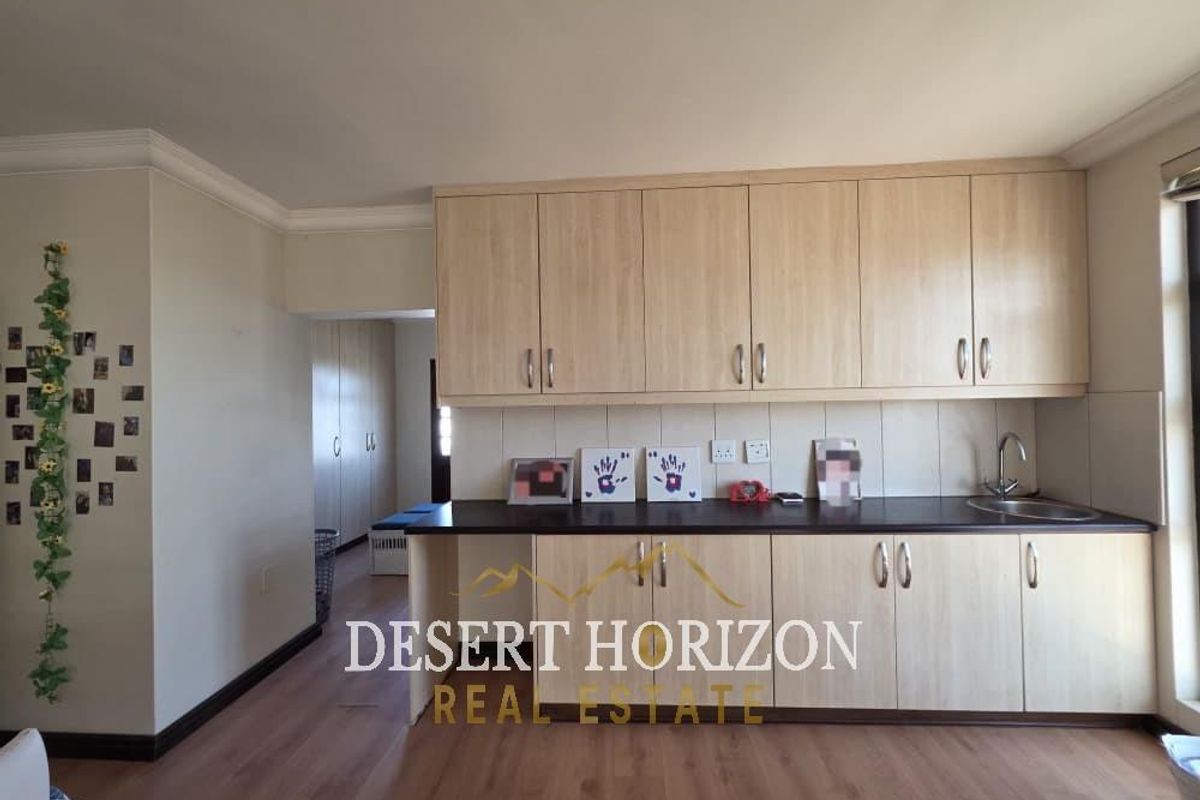 A functional kitchen area featuring light wood-finish cabinetry, dark countertops, and a stainless steel sink, set within an open-plan living space.