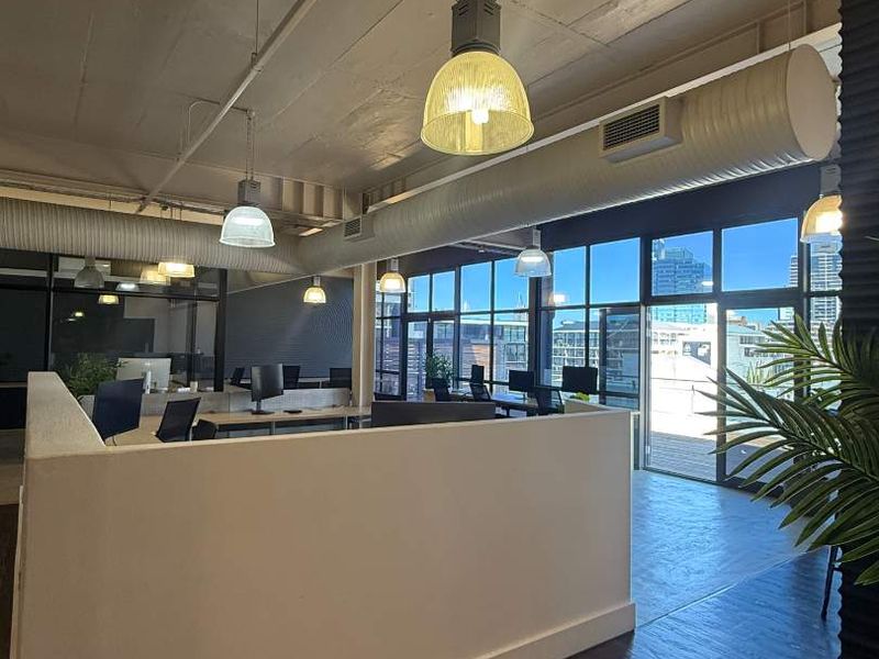 Modern open-plan office space featuring exposed concrete ceilings, industrial ducting, pendant lighting, and large windows providing ample natural light and city views.