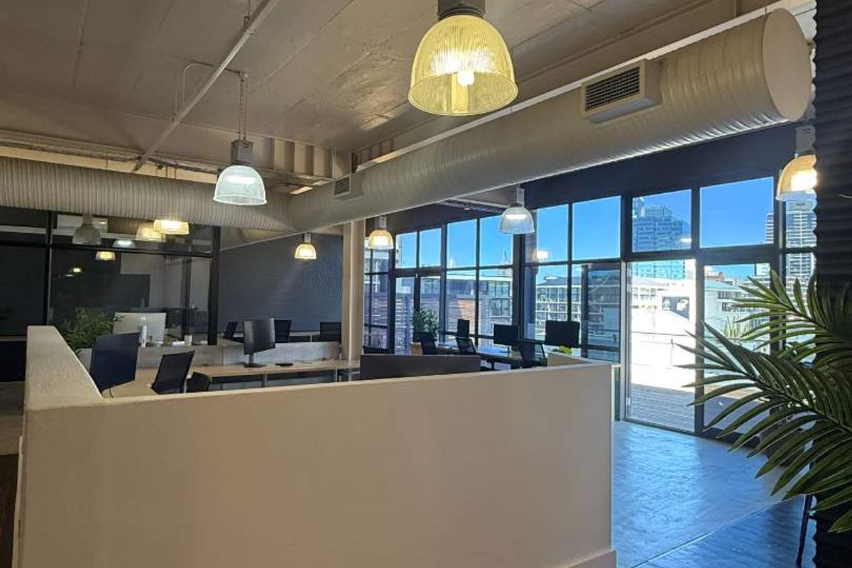 Modern open-plan office space featuring exposed concrete ceilings, industrial ducting, pendant lighting, and large windows providing ample natural light and city views.