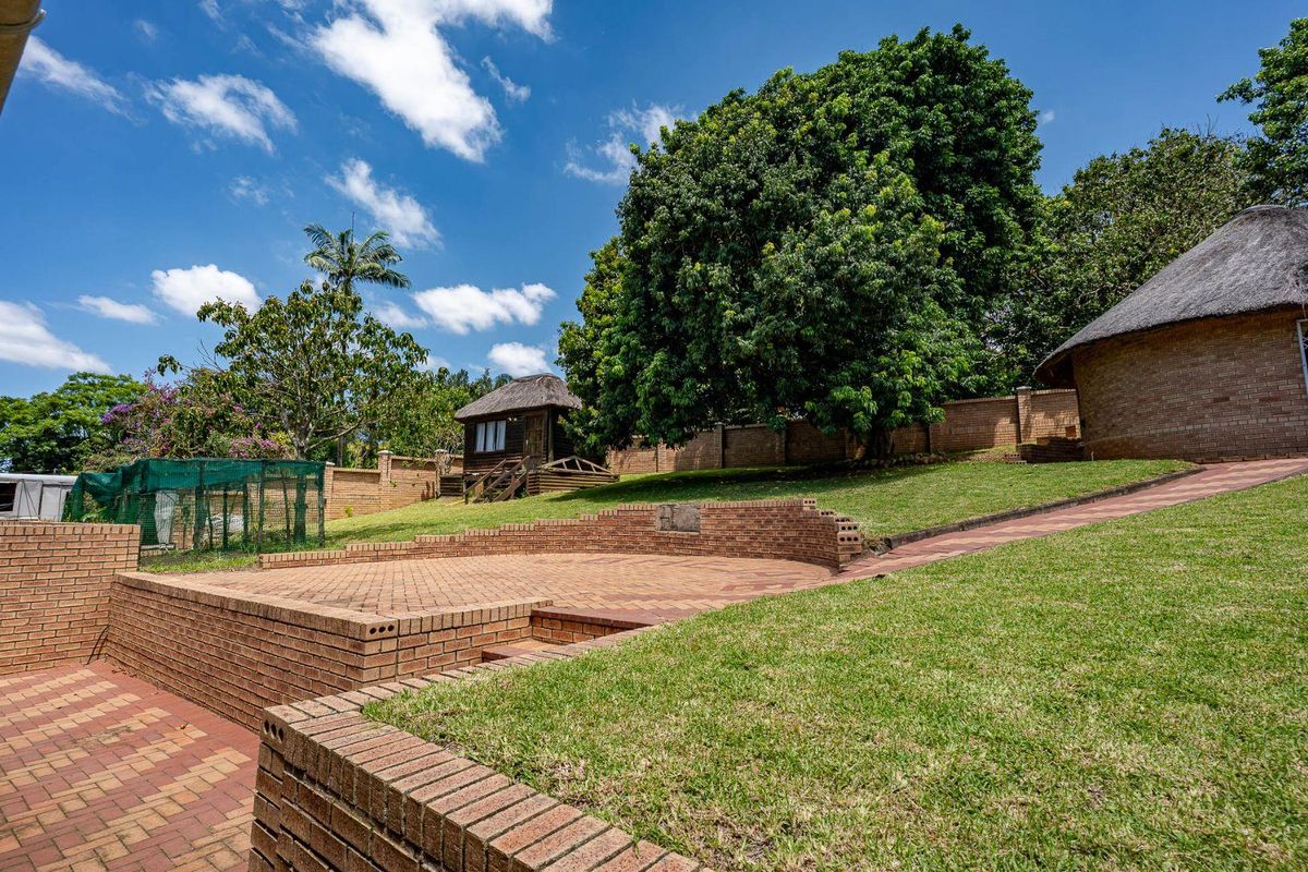 A spacious rear garden featuring tiered brick retaining walls, paved patio areas, and a well-maintained lawn with mature trees and a thatched structure.