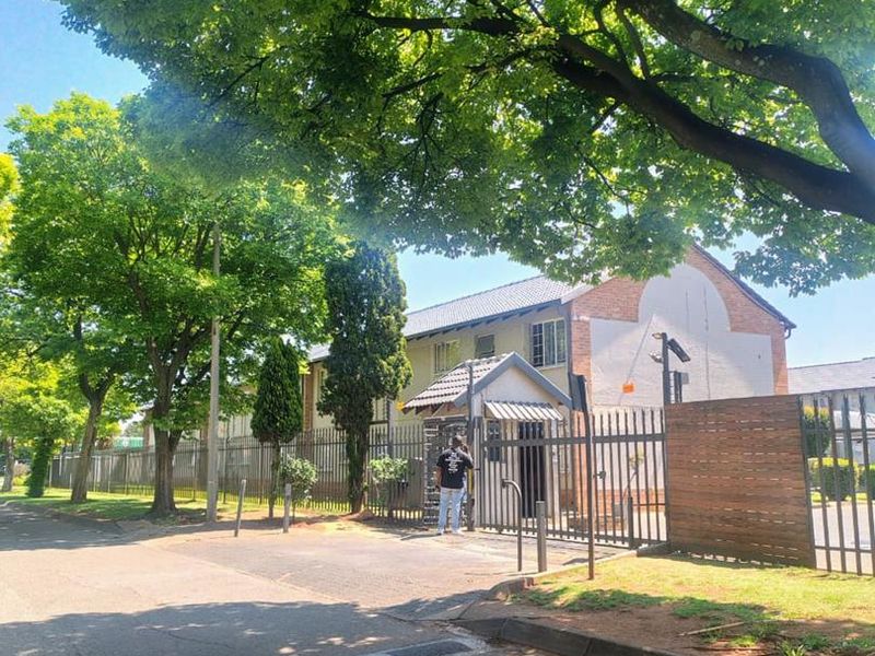 Street view of the apartment complex entrance featuring a security gate, guard house, and perimeter fencing surrounded by mature trees.
