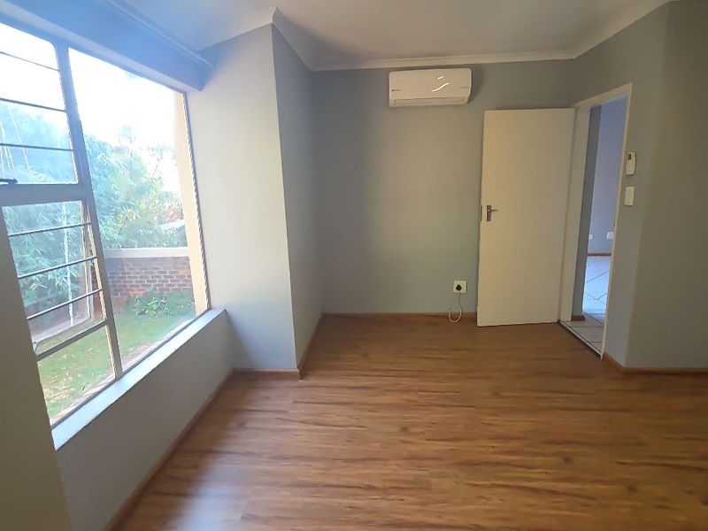 A bright, empty bedroom featuring laminate flooring, an air conditioning unit, and large windows with security bars, offering a clean and neutral living space.