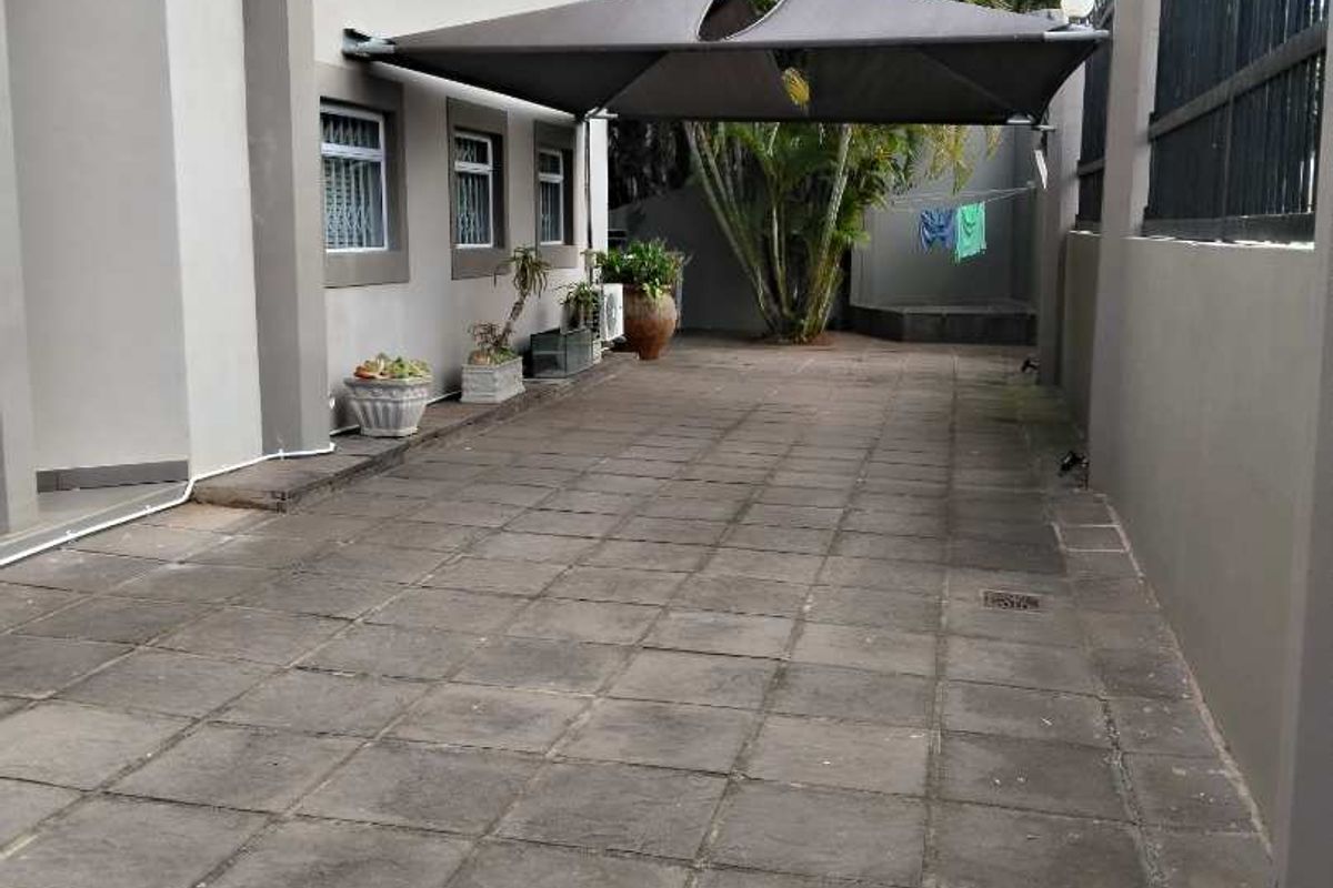 A paved rear exterior area featuring a shade sail, perimeter wall with security fencing, and exterior lighting.