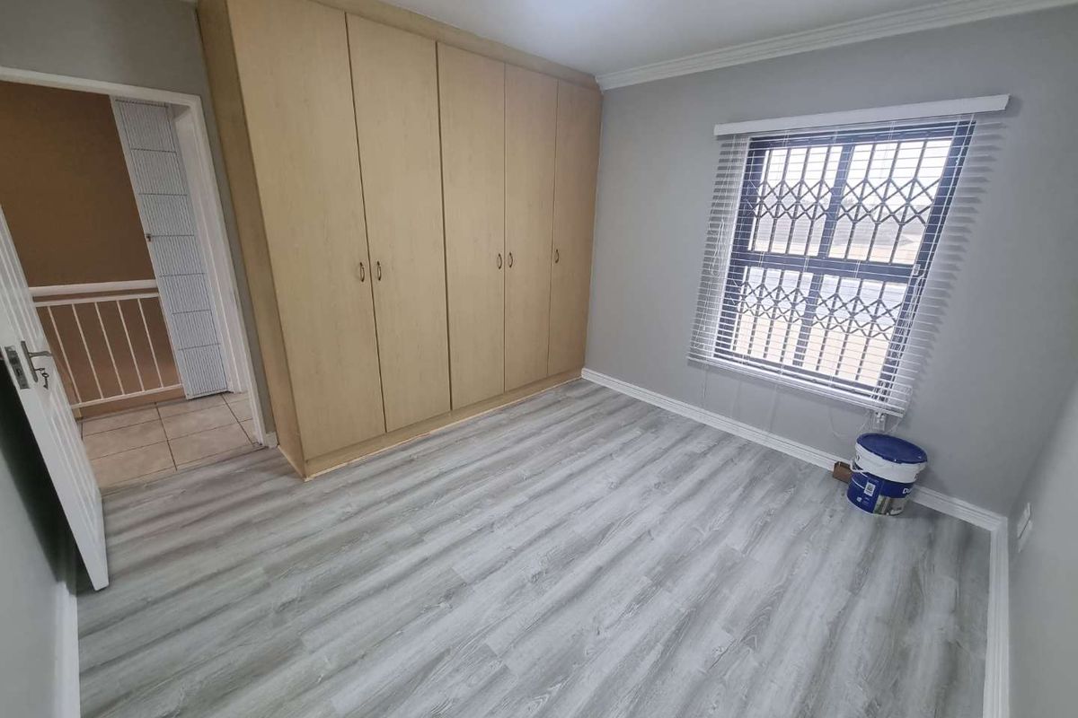 A clean, empty bedroom featuring light-toned laminate flooring, built-in wooden cupboards, and a window fitted with security bars and blinds.
