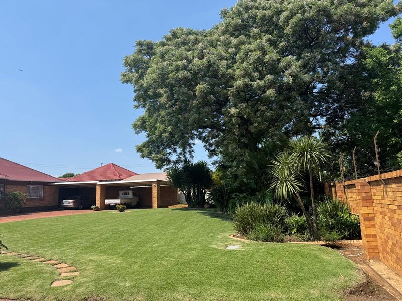 A well-maintained rear garden featuring a lush lawn, mature trees, brick boundary wall with electric fencing, and a spacious double carport.