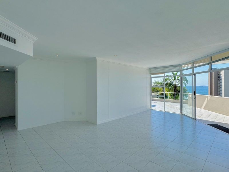 Spacious open-plan living area featuring neutral tiled flooring, ducted air conditioning, and large sliding glass doors leading to a balcony with ocean views.