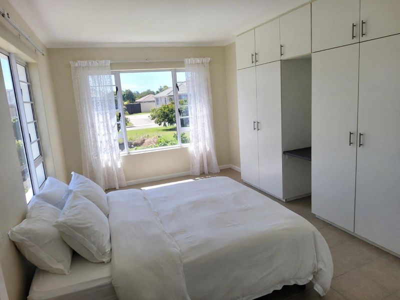 A bright bedroom featuring built-in cupboards with integrated desk space, tiled flooring, and large windows providing natural light.