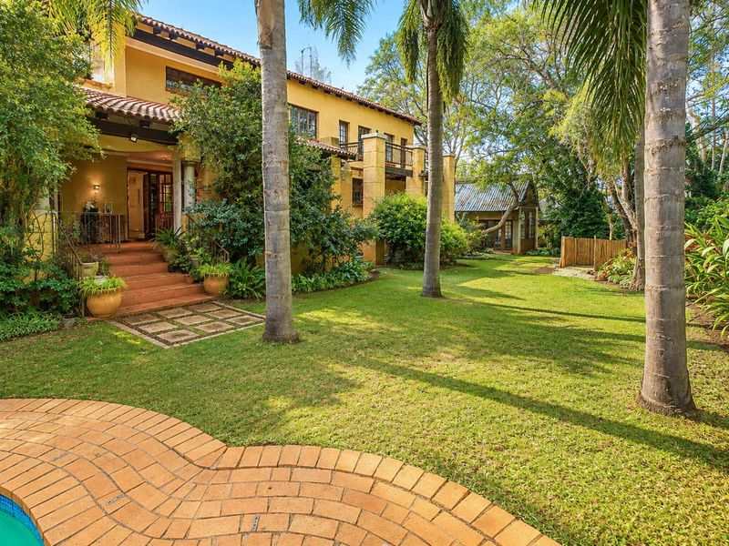 A spacious rear garden featuring a swimming pool, brick paved patio, and a multi-level residence with a balcony and terracotta roof tiles.