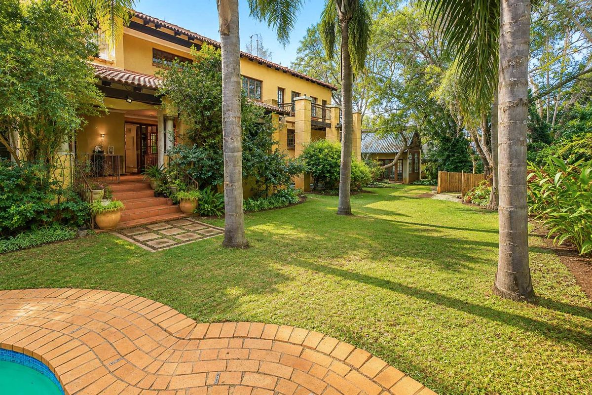A spacious rear garden featuring a swimming pool, brick paved patio, and a multi-level residence with a balcony and terracotta roof tiles.