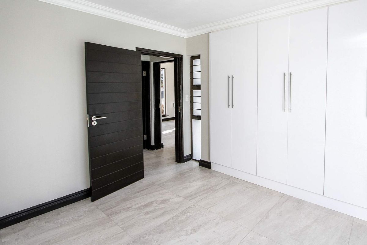 A bright, modern bedroom featuring neutral floor tiles, built-in white cabinetry, and a contrasting dark timber door leading to the hallway.