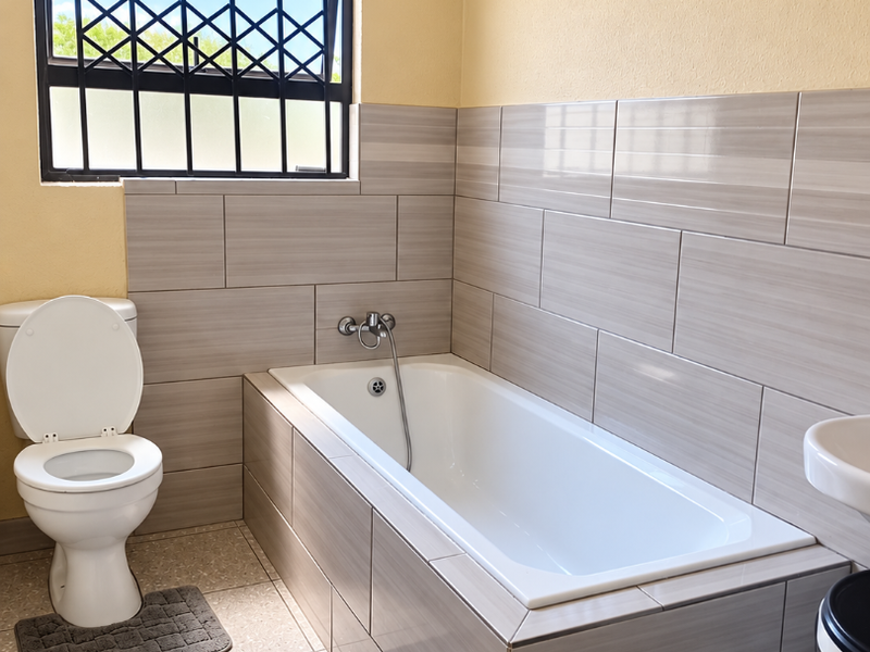 A clean, functional bathroom featuring a built-in bathtub with tiled surround, a standard toilet, and a window fitted with security bars.
