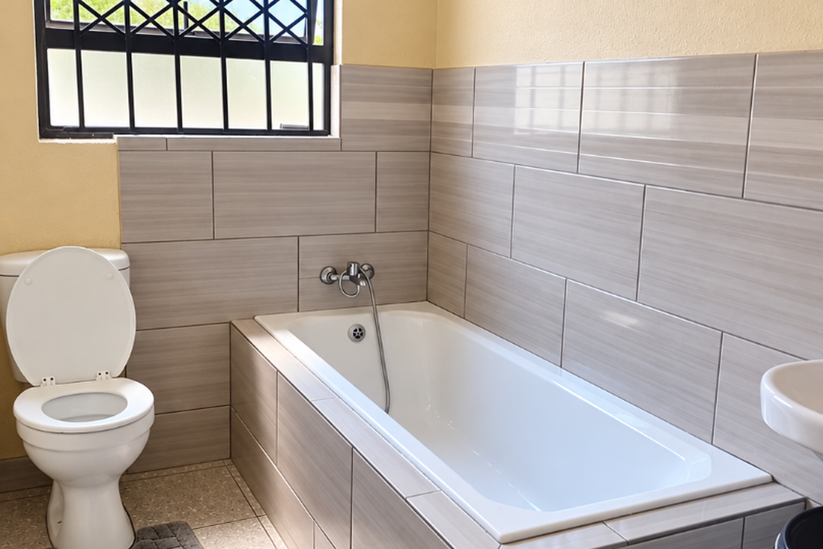 A clean, functional bathroom featuring a built-in bathtub with tiled surround, a standard toilet, and a window fitted with security bars.