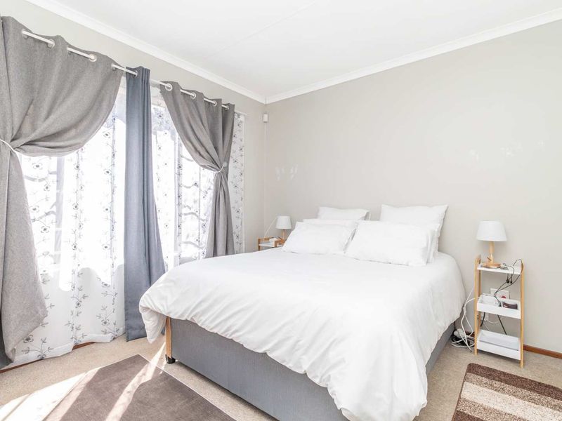 A well-lit bedroom featuring neutral wall tones, carpeted flooring, and large windows with curtain fixtures.
