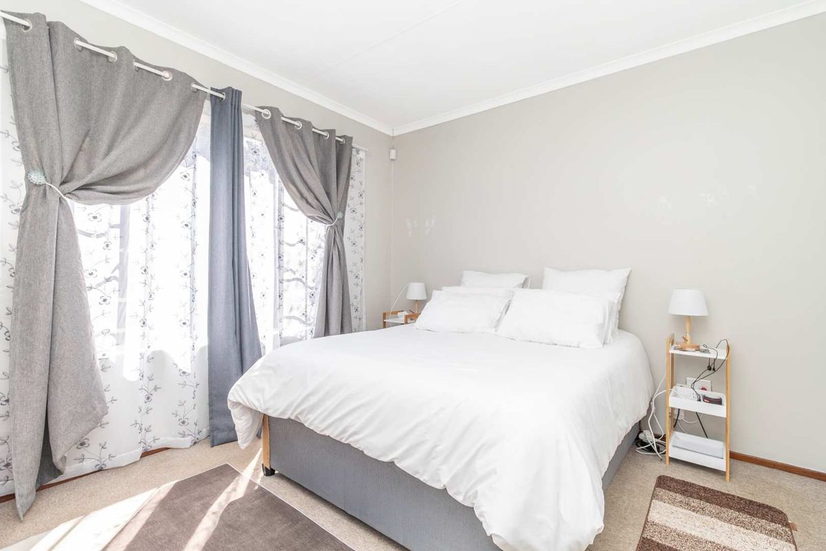 A well-lit bedroom featuring neutral wall tones, carpeted flooring, and large windows with curtain fixtures.