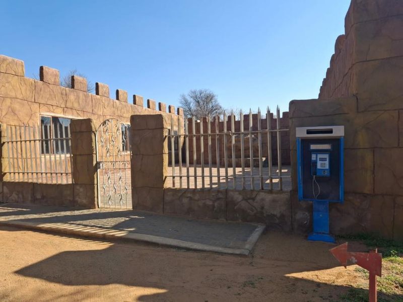 Front exterior view of the property featuring a decorative perimeter wall, metal security fencing, and a gated entrance with a public telephone booth.