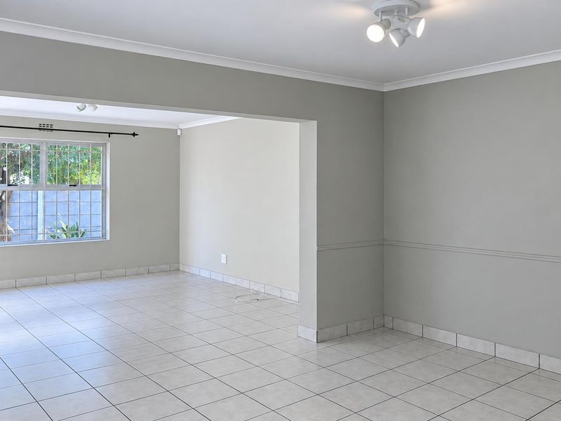 A bright, open-plan living area featuring neutral wall tones, tiled flooring, and large windows with security bars, offering a versatile blank canvas.