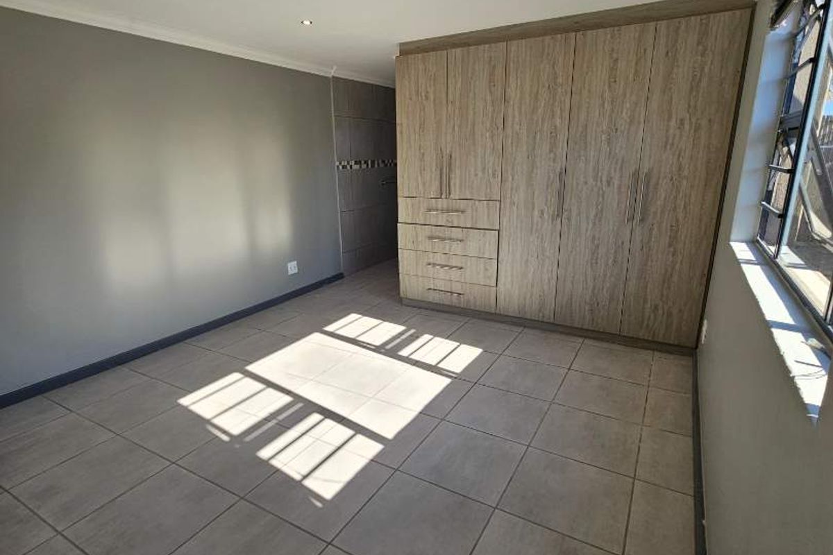 A bright, empty bedroom featuring neutral grey walls, large format floor tiles, and modern wood-finish built-in cupboards.
